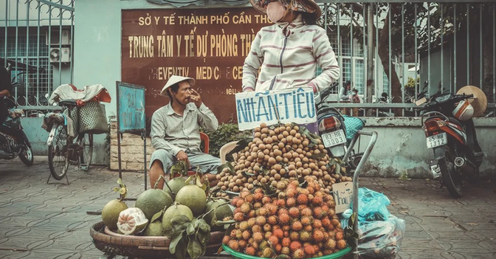 A fruit vendor in a conical hat selling pomelos and lychees on a bustling street.