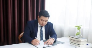 Asian businessman in a suit writing at his desk in an office environment, focusing on documents.