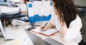 An office worker takes notes while working at a desk with a laptop and documents.