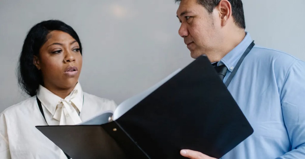 Serious middle aged male manager in shirt showing folder with documents to attentive female African American colleague during work in modern office