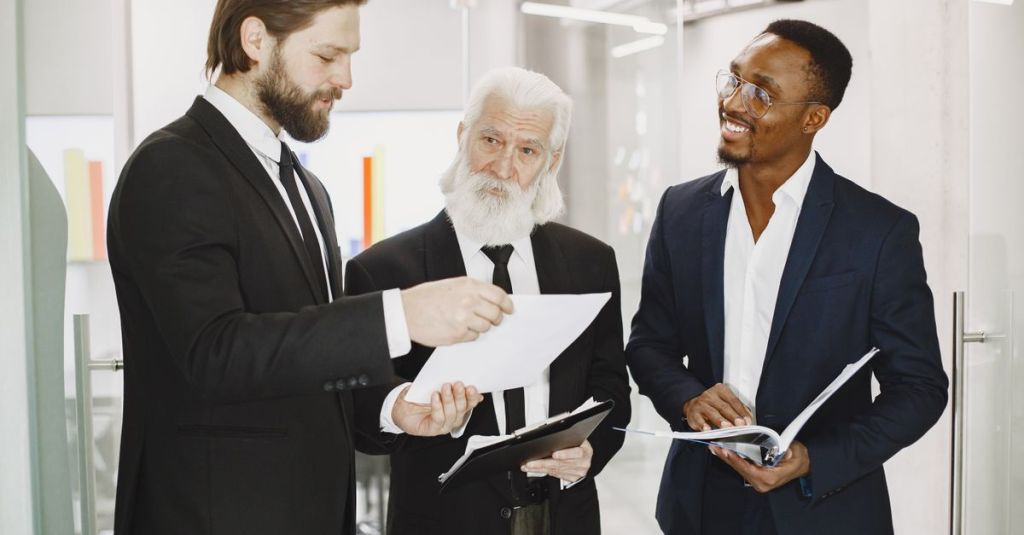 Three businessmen in suits discussing documents in a modern office setting, engaging in teamwork.