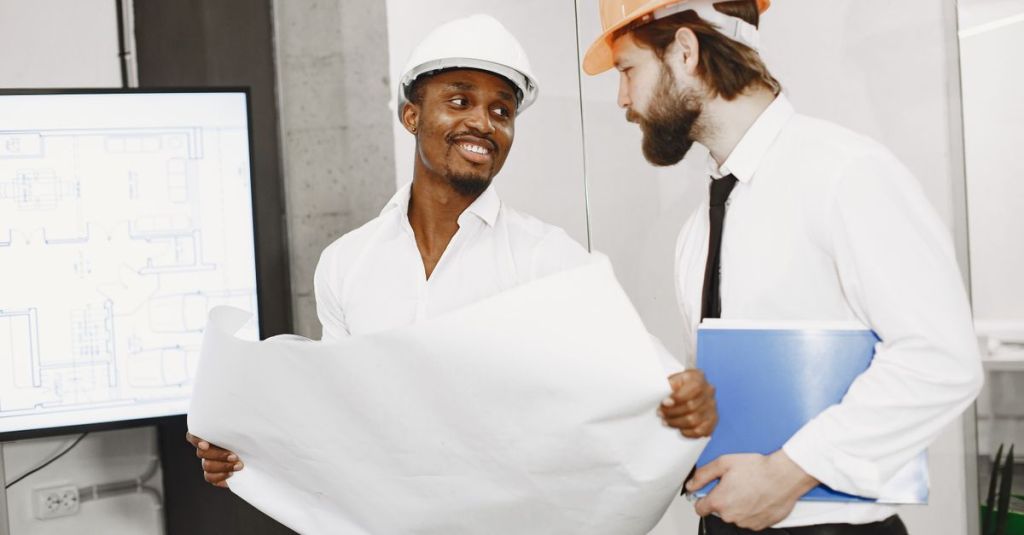 Hai kiến trúc sư đội mũ bảo hộ trao đổi về bản vẽ trong môi trường làm việc chuyên nghiệp.