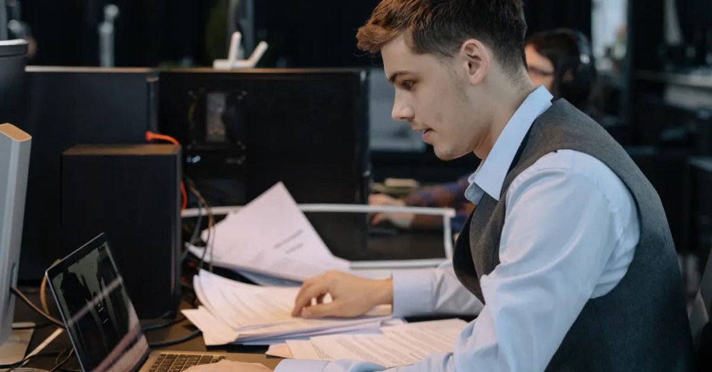 Young male professional focused on work with laptop and documents at office desk.