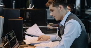 Young male professional focused on work with laptop and documents at office desk.