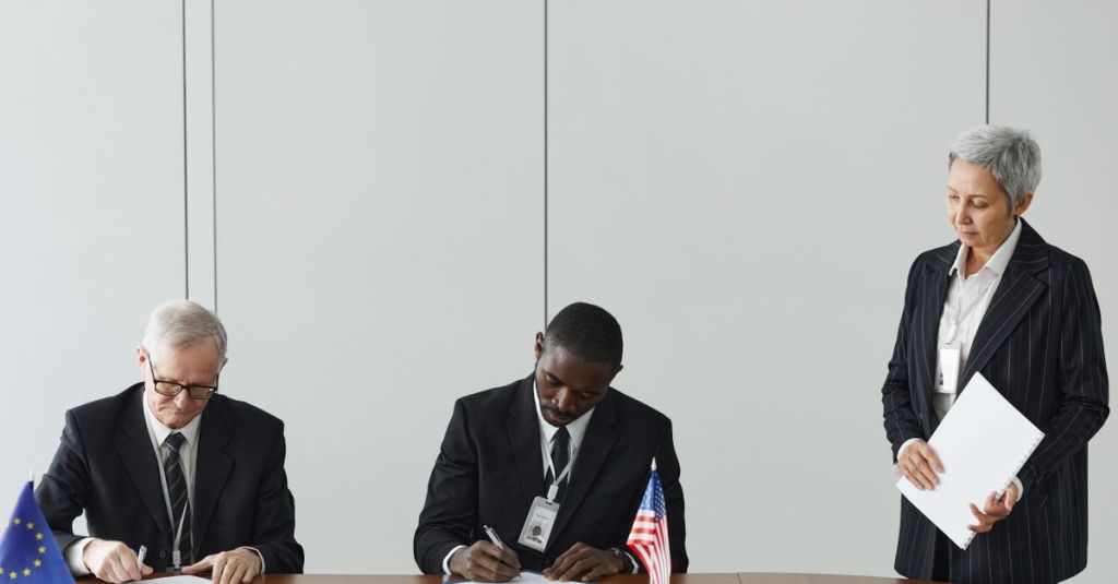 Executives signing international agreement with EU and US flags displayed on a wooden table.