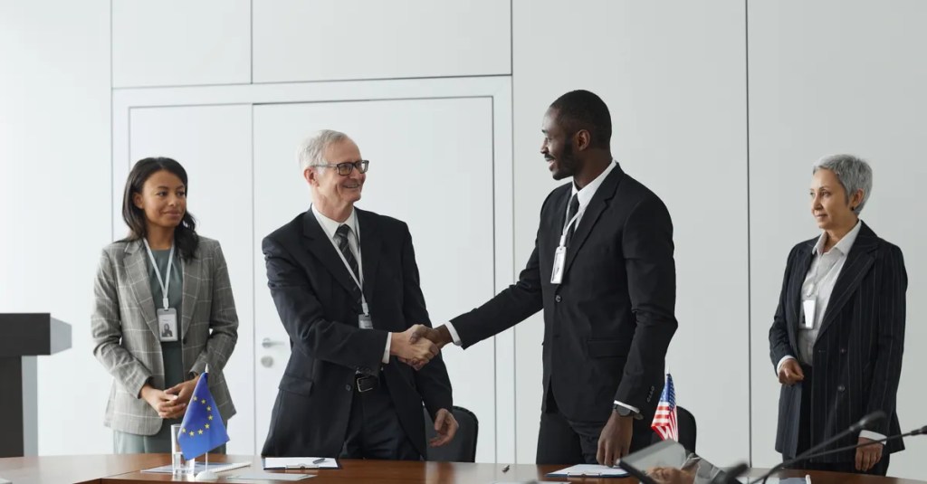 Business professionals shaking hands in conference room with flags signifying international agreement.