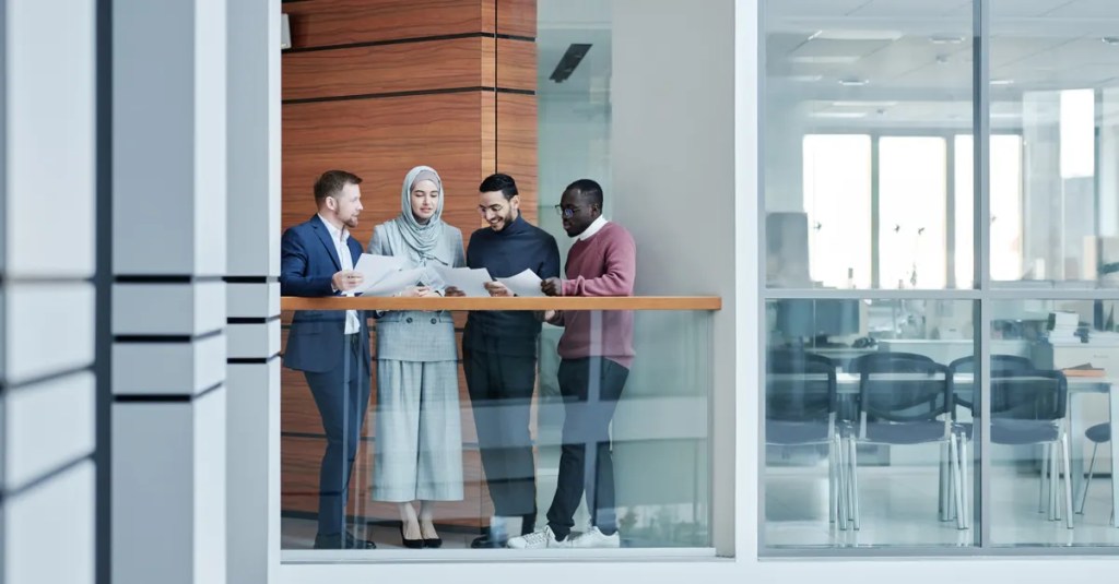 Group of diverse colleagues collaborating in a modern office with glass walls, emphasizing teamwork and inclusivity.
