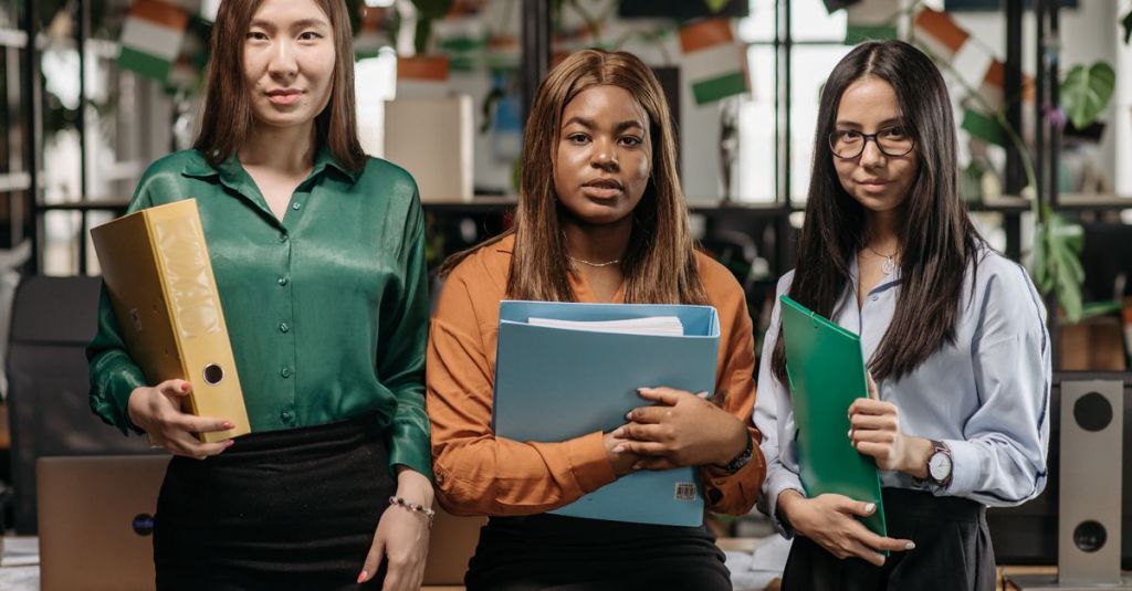 Three diverse women holding binders in an office, representing teamwork and diversity.