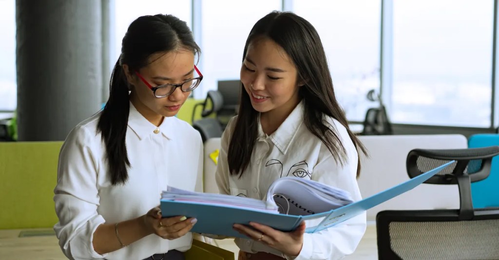 Two professional women reviewing documents together in a modern office setting, emphasizing teamwork.