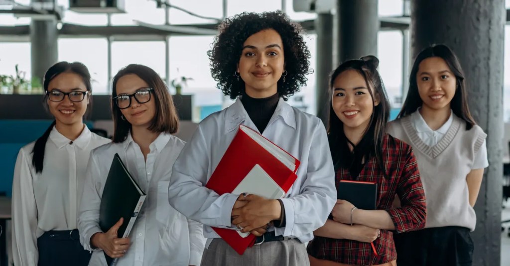 Group of diverse women colleagues smiling in a modern office setting, holding documents.
