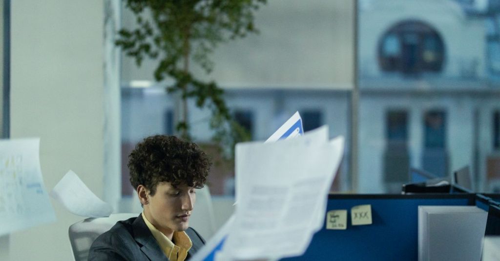 Young man in office cubicle tossing papers, illustrating busy work environment.